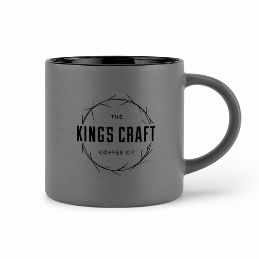 A dark gray ceramic coffee mug with a black interior and handle, featuring a black printed logo for 'The King's Craft Coffee Co' in the center, set against a plain white background.
